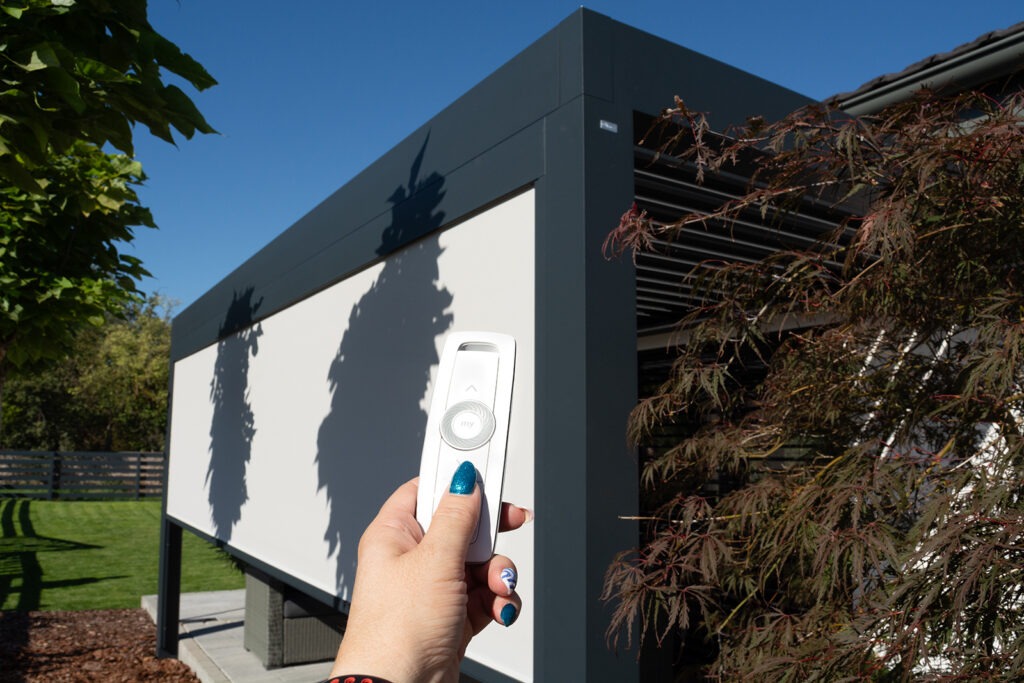 Dálkový ovladač v ruce před pergolou se spuštěnou screenovou clonou